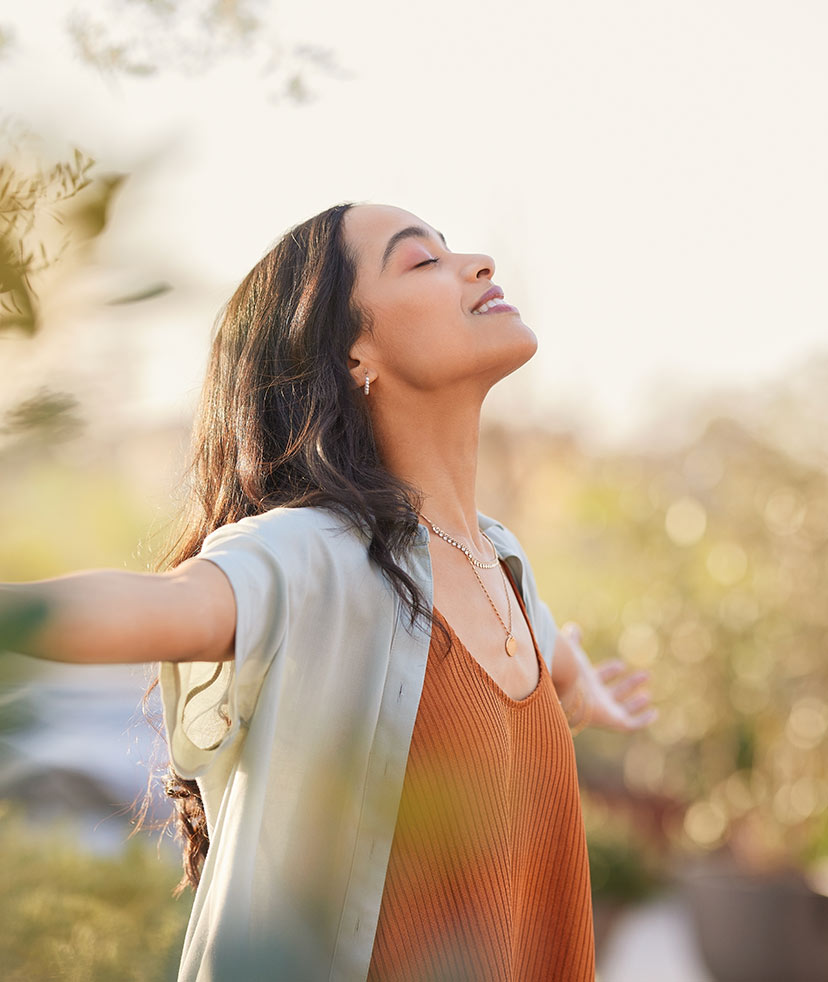 A woman with long dark hair stands outdoors with her arms outstretched, eyes closed, and a peaceful smile on her face, enjoying the sunlight and fresh air.