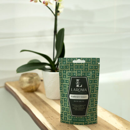 A green packet of Dream Haven Bath Salts (CBD Isolates) rests on a wooden bath tray next to a potted orchid and a small gold candle holder, with a white tiled wall in the background.