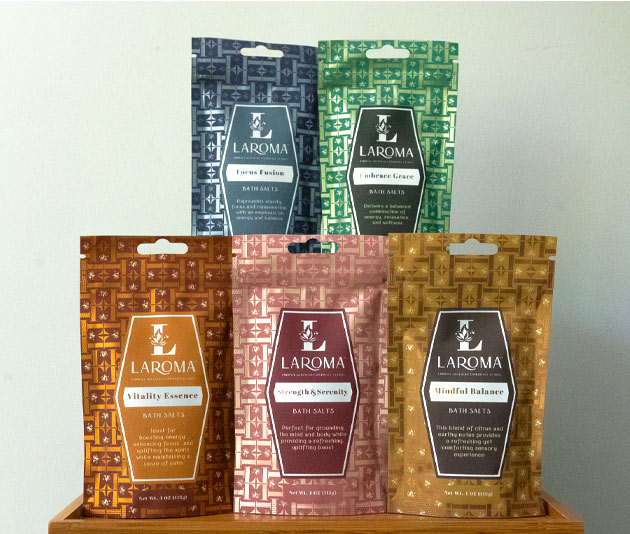 Six colorful Laroma bath salts packets, each with distinct patterns and labels, are displayed upright on a wooden shelf against a plain background.