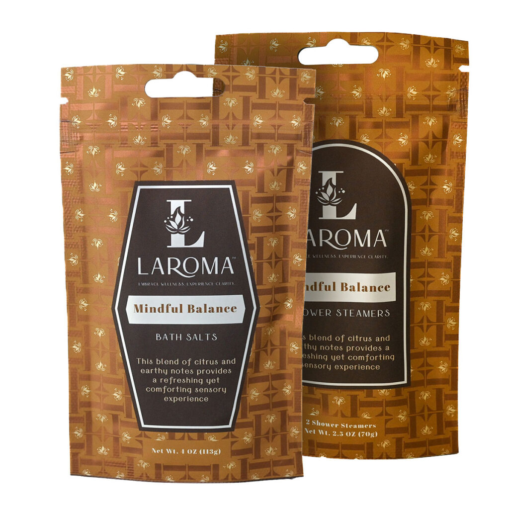 Two brown packets of LAROMA Mindful Balance products are shown standing upright. One contains bath salts, and the other contains shower steamers. Both have geometric patterns and product descriptions on the front labels.