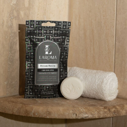 A package of Embrace Grace Shower Steamers sits on a tiled shower shelf beside a neatly rolled white towel and a single round shower steamer.