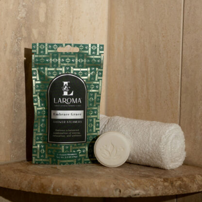 A green Embrace Grace Shower Steamers box sits on a stone ledge beside a rolled white towel and a round floral soap, set against a beige stone backdrop.