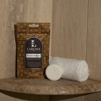 An Embrace Grace Shower Steamers pouch, a white rolled towel, and a round embossed shower steamer sit on a stone shower ledge against a beige tiled wall.