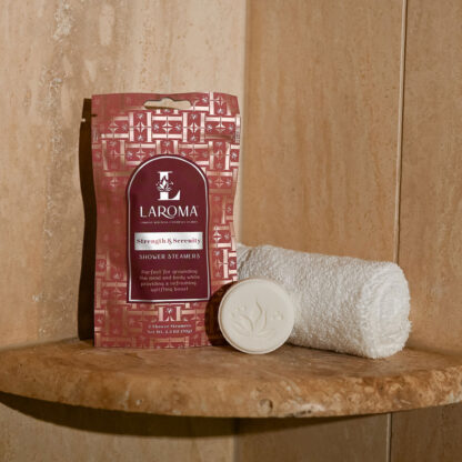 An Embrace Grace Shower Steamers package stands upright on a stone ledge beside a rolled white towel and a round white soap, set in a beige tiled bathroom.