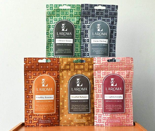 Five colorful Laroma shower steamer packages—green, blue, orange, yellow, and red—are displayed upright on a wooden surface, each labeled with different aromas and benefits.