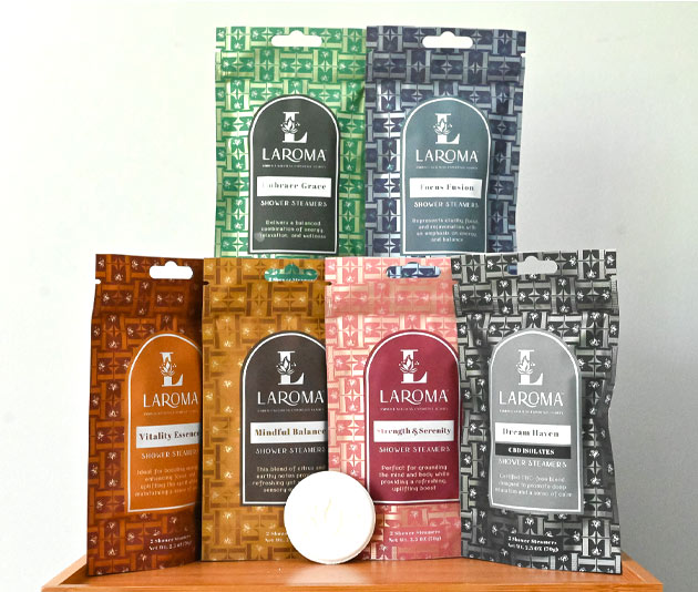 Six colorful LAROMA branded bath product pouches are arranged in two rows on a wooden surface, with a white round bath bomb in front of them. Each pouch features distinct colors and labeled scent names.