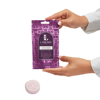 Two hands hold a purple Mindful Balance Shower Steamers package, with a round, pale pink bath tablet featuring a leaf design placed below it on a white background.