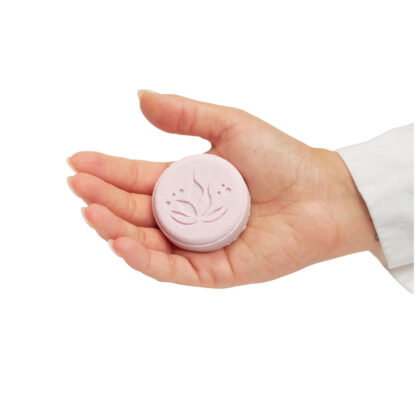 A hand holds a round, light pink Mindful Balance Shower Steamer with a flower design embossed on its surface, set against a white background.