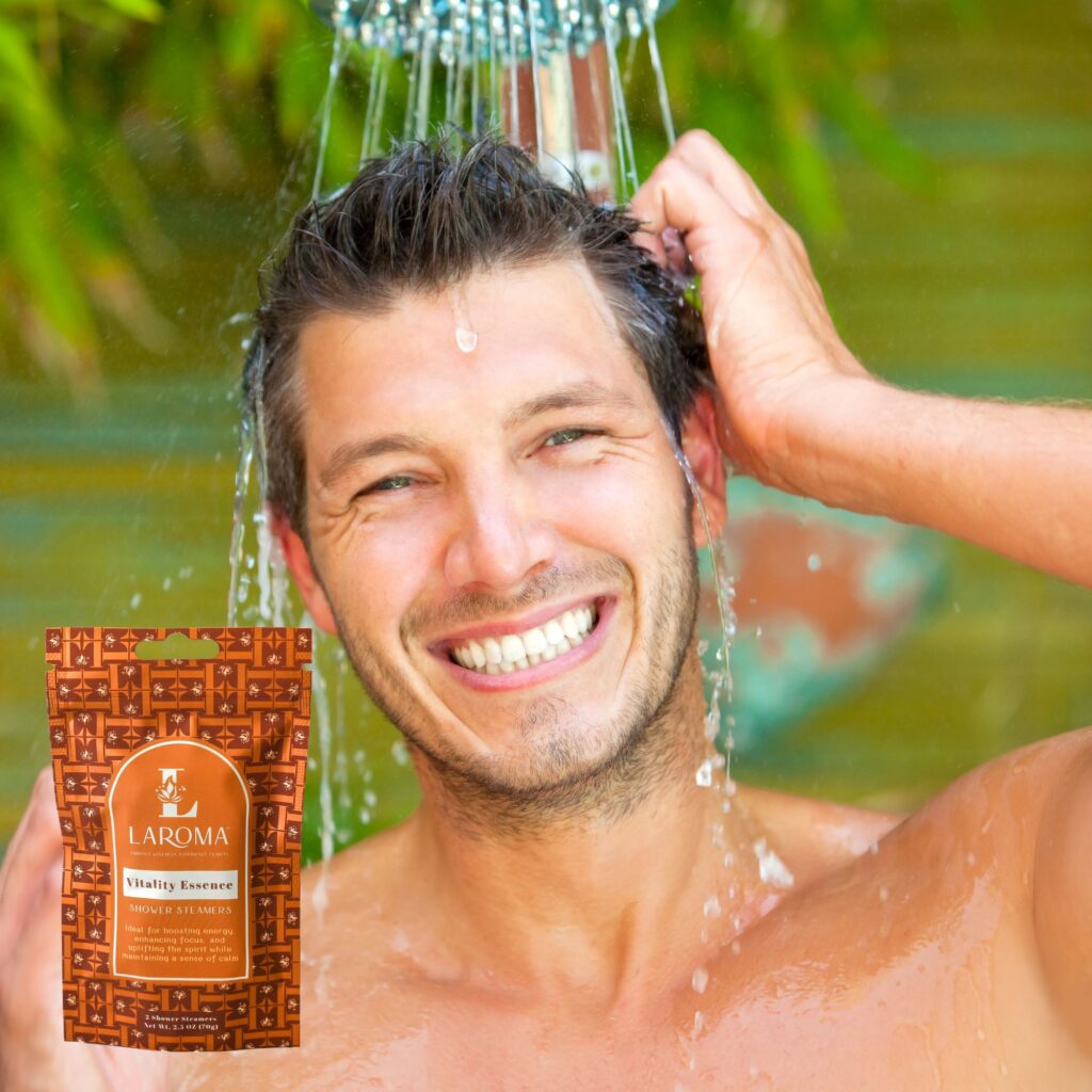 A smiling man stands under a shower outdoors, rinsing his hair. In the foreground, a brown packet labeled LAROMA Vitality Essence Shampoo & Sulfate Free is visible. Green foliage is blurred in the background.