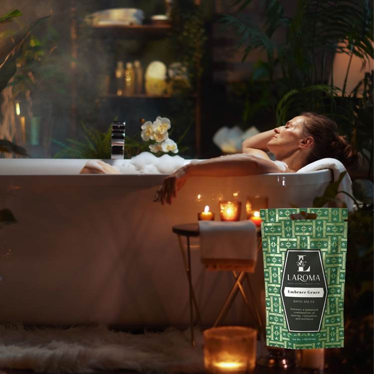A woman relaxes in a bathtub surrounded by candles and plants in a dimly lit room. In the foreground, a green package labeled LAROMA Bamboo Grove Bath Salt Soak is prominently displayed.